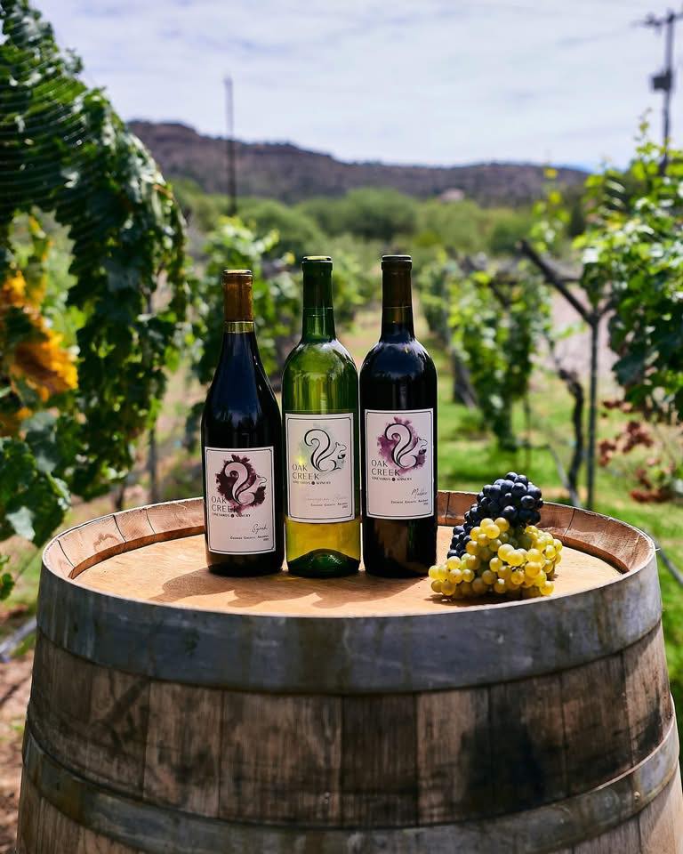 Oak Creek Vineyards and Winery Three Oak Creek Vineyards wine bottles on barrel with grapes in vineyard landscape.